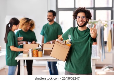 charity, donation and volunteering concept - happy smiling male volunteer with food in box pointing to camera over international group of people at distribution or refugee assistance center