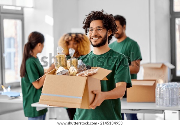 charity, donation and\
volunteering concept - happy smiling male volunteer with food in\
box and international group of people at distribution or refugee\
assistance center