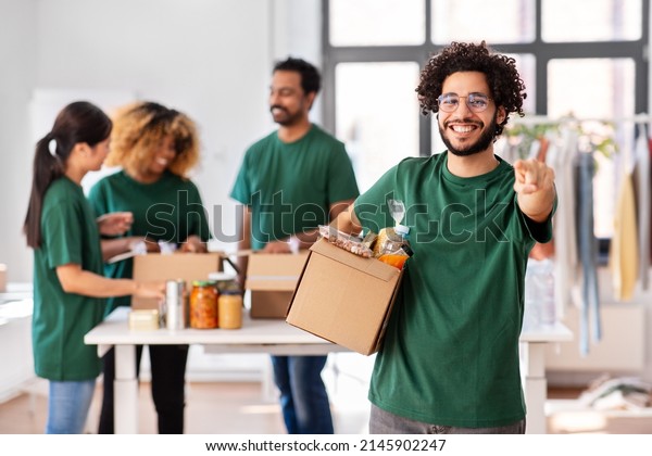 charity, donation and volunteering concept - happy\
smiling male volunteer with food in box pointing to camera over\
international group of people at distribution or refugee assistance\
center