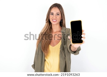 Charming adorable young beautiful woman wearing green overshirt over white background holding modern device, showing black screen smartphone