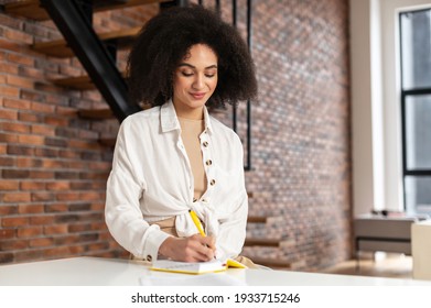 Charming African American Girl With Curly Dark Hair Writing Down In The Notebook Thoughts, Phone Number, Resolutions, Making A Plans For A Day, Dressed In Casual Style, Holding A Pen In The Hand