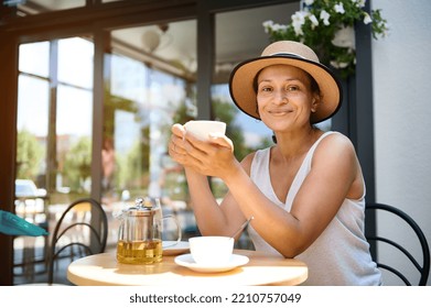 Charming African American woman in a straw hat, smiling a beautiful toothy smile, looking at camera, holding a cup of hot drink and enjoying her coffee break in cozy summer terrace. Tea or coffee time