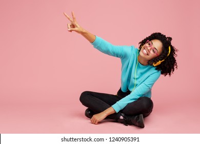 Charming African American Young Woman With Bright Smile Dressed In Casual Clothes, Glasses And Headphones Showing Peace Gesture Over Pink Background.