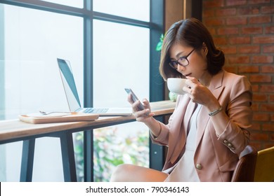 Charming Asian Business Woman On Pink Suit Drinking Coffee And Using Mobile Phone In The Coffee Cafe.