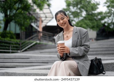 A charming Asian businesswoman in a business suit is sitting on stairs outdoor in the city, holding a takeaway coffee cup and smiling at the camera. - Powered by Shutterstock