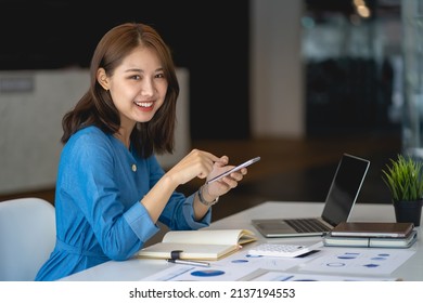 Charming Asian woman with a smile sitting holding smartphone at the office. - Shutterstock ID 2137194553