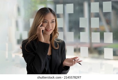 Charming Asian Woman With A Smile Standing Holding Pen And Mobile Phone At The Office.
