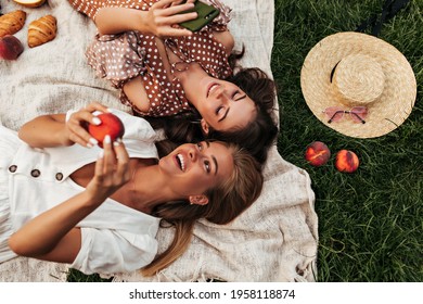 Charming attractive blonde and brunette girls in summer dresses lay on linen rug and have picnic. Cool woman in polka dot dress holds phone.