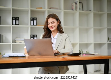 Charming And Attractive Young Caucasian Businesswoman Or Female Marketing Manager In Business Suit Looking At Her Laptop Screen, Using Notebook Laptop, Working In Her Modern Office.