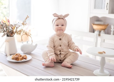 A Charming Baby Girl In A Jumpsuit Made Of Natural Fabric Is Sitting On The Table At Home In A Bright Kitchen, The First Lure