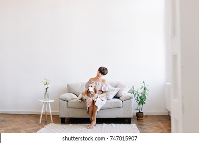 Charming barefooted girl sitting on gray sofa between little table and green plant, looking at beagle dog on her knees. Pensive brunette woman resting with puppy in big light room.