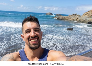 Charming Bearded Latino Sharing Selfie In Social Media In The Beach With Close Eyes Because Of The Sun. Tan Fit Muscle Surfer With Tattoo In The Arm Showing Teeth, Charming And Smiling In The Ocean