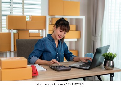 Charming beautiful Asian teenager owner business woman work at home for online shopping, calculate price of goods with laptop with office equipment, entrepreneur lifestyle concept