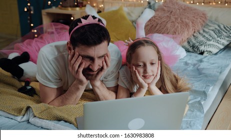 Charming beautiful fairies dad and daughter watching exciting cartoons on laptop resting on bed having a custom disney party at home.