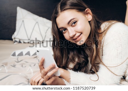charming beautiful girl with clean skin lies in bed, holds a smartphone in her hand and smiles