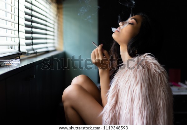 魅力的な美しいセクシーな女性がマリファナかタバコを吸っている 魅力的な美しい女の子は リラックスして幸せを感じます 彼女はぼんやりしている あるいは夢のような状態にある 美しいアジア人の女性は毛皮のコートを着る の写真素材 今すぐ編集