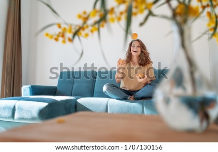 Charming beautiful young girl with curly hair and a happy smile juggles tangerines while sitting on a blue sofa in her living room at home, having fun staying at home