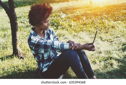 Charming Black Female Teen With Digital Tablet Sitting In Park On Summertime And Reading EBook, Attractive Mixed Girl With Curly Afro Hair And Touch Pad Sitting On Grass Of Meadow On Spring Sunny Day