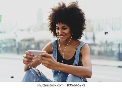 Charming Black Girl With A Charming Smile Book Tickets Online Via Cellphone.a Young Woman With Beautiful Afro Hair Prints Email Using A Smartphone.Female Person Listening Music Via Headphones Outside