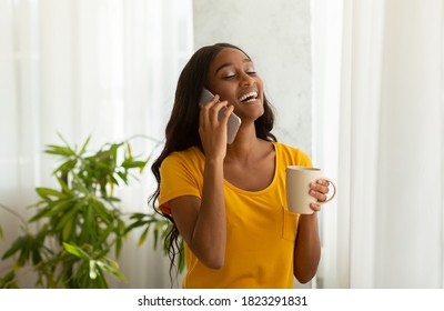 Charming black woman speaking on cellphone with cup of aromatic coffee at home. Attractive African American lady having conversation on mobile phone near window indoors