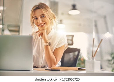 テーブルに座りながらカメラを見て微笑み、仕事にノートパソコンを持つ魅力的な金髪の女性