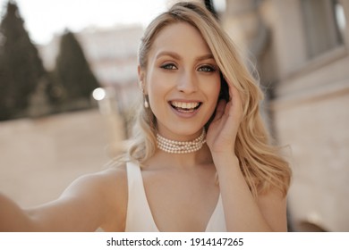 Charming blonde woman in pearl necklace and white top takes selfie and smiles sincerely at street.