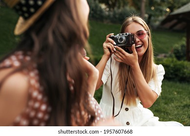 Charming blonde woman in stylish white dress and red sunglasses smiles and takes photo of her friend. Tanned girl holds retro camera. Brunette lady in boater has picnic. Blurred effect. - Powered by Shutterstock