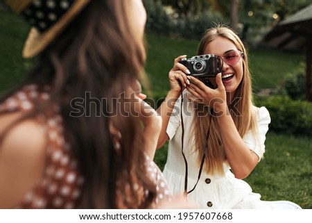 Charming blonde woman in stylish white dress and red sunglasses smiles and takes photo of her friend. Tanned girl holds retro camera. Brunette lady in boater has picnic. Blurred effect.