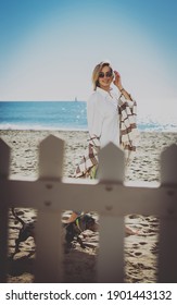 Charming Blonde Woman Wearing Sunglasses And Casual Outfit Looking At The Camera While Walking With The Pet Dog Behind The Fence On The Beach. Girl Enjoying The Day With Her Puppy On A Sandy Beach.