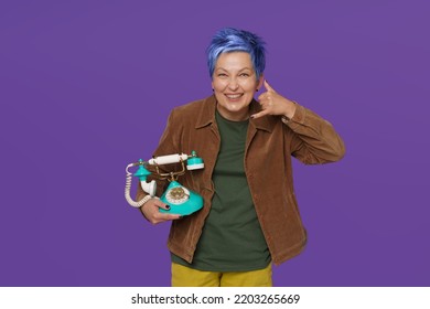 Charming Blue Haired Senior Woman Hold Vintage, Retro Phone And Gesturing CALL ME Or Phone Flirt Wearing Casual Isolated On Purple Background. Short Haired Lady With Old Phone In Hands. 