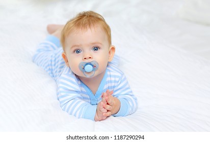 Charming blue-eyed baby 7 month old lies in bed in a striped bodysuit and sucks a pacifier