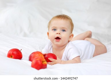 Charming blue-eyed baby 7 months lying in bed in white clothes with red apples
