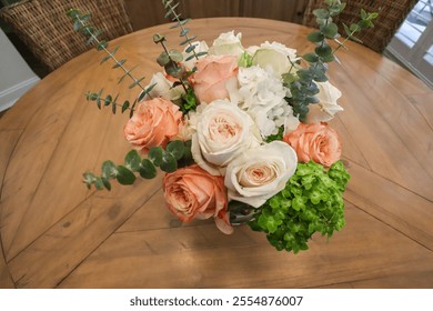 A charming bouquet of peach-orange and white roses complemented by fresh eucalyptus, soft green hydrangea, and delicate greenery, arranged elegantly in a glass vase on a wooden table. - Powered by Shutterstock