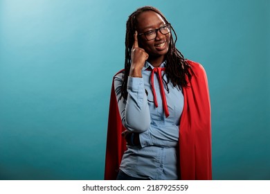 Charming And Brave Young Superhero Woman Wearing Justice Defender Hero Cape While Pointing Finger At Temple. Strong And Joyful Adult Person With Superpower Abiliies Smiling Heartily At Camera.