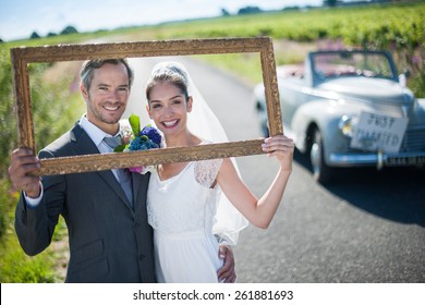 Charming Bride And Groom Posing With A Frame For Their Wedding Photos, Behind Them A Lovely Country Road