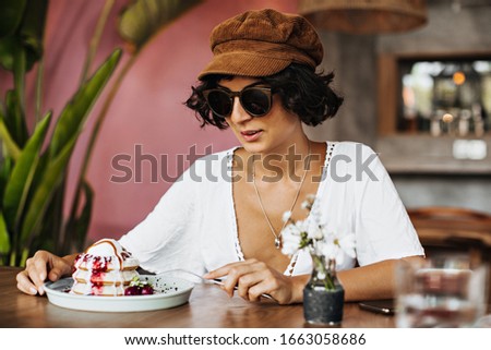 Charming brunette tanned woman in sunglasses and cap eats dessert in cafe. Girl enjoys fluffy pancakes with ice cream and strawberry sauce