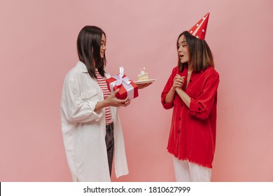 Charming Brunette Tanned Woman In White Oversized Blouse Smiles, Presents Tasty Birthday Cake And Red Gift Box To Her Friend. Young Asian Girl Celebrating B-day On Pink Background.