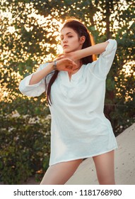 A Charming Brunette In A White Long Shirt With Bare Legs Poses Standing On The Sand In The Warm Summer Day. The Golden Hour Of The Rays Of The Sun Is Visible Everywhere In The Leaves Of The Tree