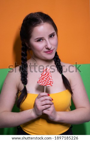 charming brunette woman in yellow top holding red big lollipop on bright green and orange background. sweets concept. candy. copy space