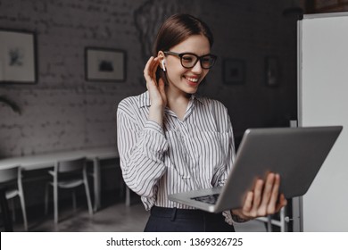 Charming business woman in headphones and glasses is laughing, looking into laptop screen against background of office