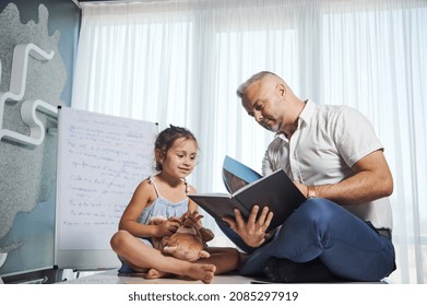 Charming Businessman, Successful Entrepreneur, Loving Father Sits On The Table Next To His Cute Little Daughter, Shows Her A Book, Reads Fairy Tales, Pays Attention To Her Despite A Busy Work Schedule