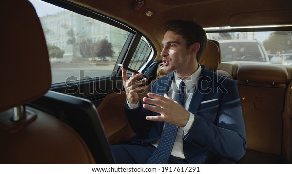 Charming businessman telling story in voice message\
to smartphone in modern car. Smiling male professional making voice\
recording in business car. Excited business man sharing news on\
phone in car.