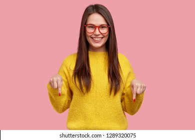 Charming carefree woman with positive expression, points down with both index fingers, dressed in casual clothing, has broad intrested smile, isolated over pink background. Look there, please