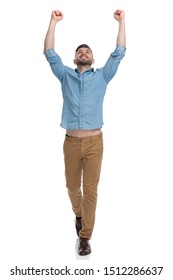 Charming Casual Man With Blue Shirt Walking With Hands Up In The Air While Looking Up Victorious On White Studio Background