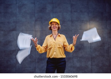 Charming Caucasian Blonde Senior Female Architect With Short Hair And Helmet On Head Standing Outdoors In Front Of Wall And Throwing In The Air Papers.