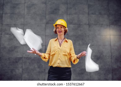 Charming Caucasian Blonde Senior Female Architect With Short Hair And Helmet On Head Standing Outdoors In Front Of Wall And Throwing In The Air Papers.