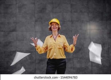 Charming Caucasian Blonde Senior Female Architect With Short Hair And Helmet On Head Standing Outdoors In Front Of Wall And Throwing In The Air Papers.