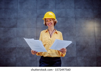 Charming Caucasian Blonde Senior Female Architect With Short Hair And Helmet On Head Standing Outdoors In Front Of Wall And Holding Important Documents While Looking At Camera.