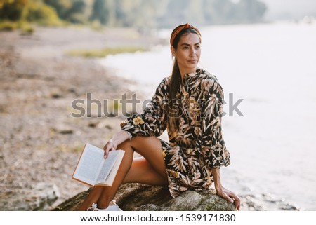 Charming caucasian brunette with headband and in floral dress sitting on rock near river, holding book and looking away.