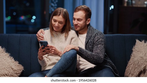 Charming Caucasian Couple Staying Home Using Mobile Phone Together. Affectionate Young Happy Girlfriend And Boyfirend Dating In Apartment At Night.